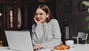 Cheerful girl in gray outfit working in laptop during lunch with croissant and cup of coffee.