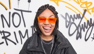 Cheerful fancy teenage girl wears trendy orange suglasses and black jacket sticks out tongue has dreadlocks hairstyle expresses positive emotions poses against graffiti wall. Swag and style concept