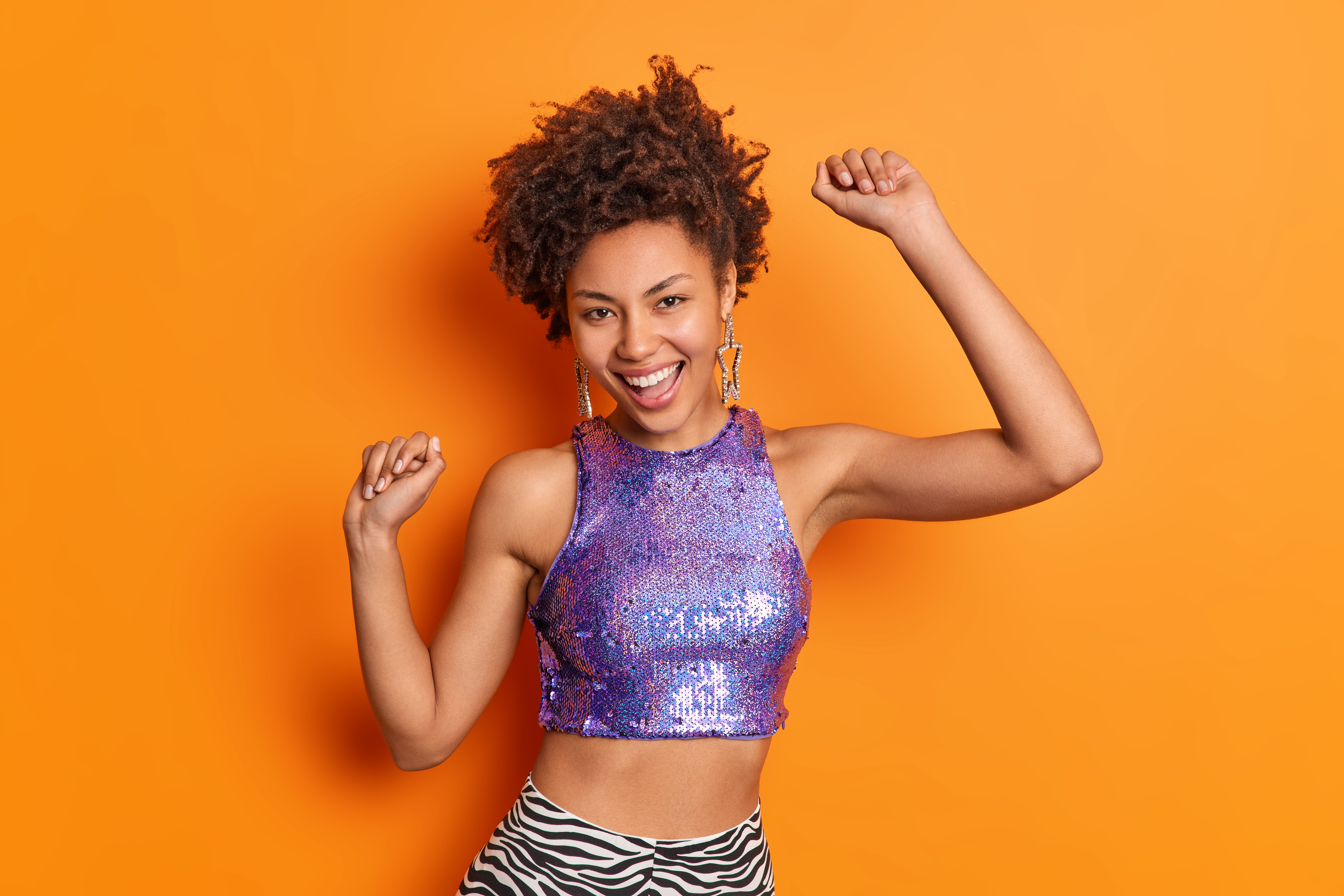 Glad dark skinned fashionable Afro American woman with curly hair dances carefree dressed in purple glittering top raises arms isolated over vivid orange background. People mood fashion concept