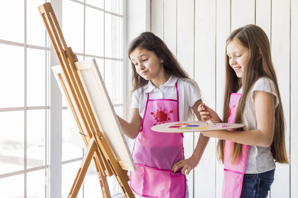 close-up-two-girls-standing-near-window-painting-easel-with-paint-brush