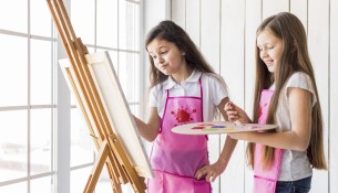 close-up-two-girls-standing-near-window-painting-easel-with-paint-brush