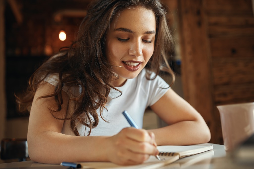 Close up of charming plus size teenage girl with loose wavy hair sitting at desk with notebook, handwriting, drawing or making sketches, having joyful look. Creativity, hobby and leisure concept