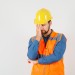 Young builder standing in thinking pose in shirt, vest, helmet and looking tired , front view.