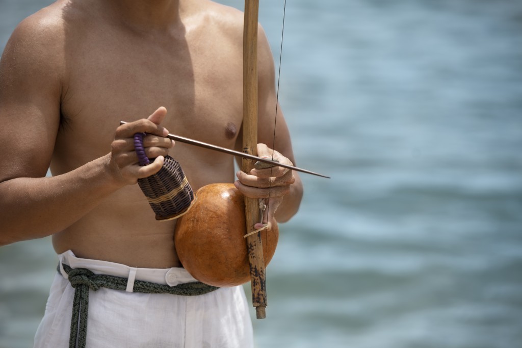 shirtless-man-practicing-capoeira-by-beach-with-wooden-bow