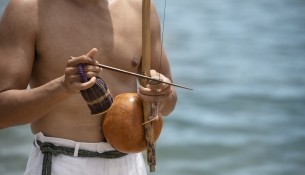 shirtless-man-practicing-capoeira-by-beach-with-wooden-bow