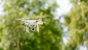 White drone in forest. Copter with digital camera flying high in the air, taking photos, recording footage. Drone against the forest background. Free space.