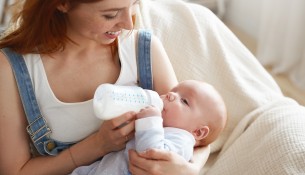 Sweet moment of happy young Caucasian mother smiling joyfully holding bottle of modified milk and feeding her adorable baby. People, lifestyle, childcare, maternity, love, family and happiness concept