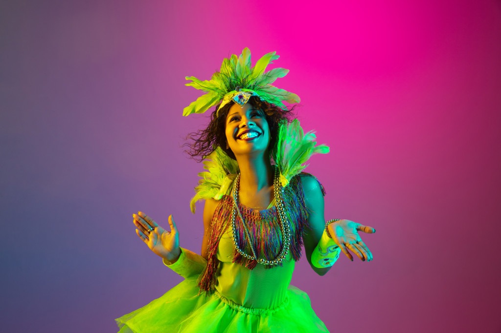 Beautiful young woman in carnival and masquerade costume on gradient studio background in neon light