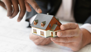 Close up shot of Caucasian contractor holding real estate project while his African colleague pointing finger at scale model building, explaining design during presentation meeting in office