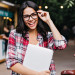 Blissful dark-haired girl with laptop touching her glasses. Outd